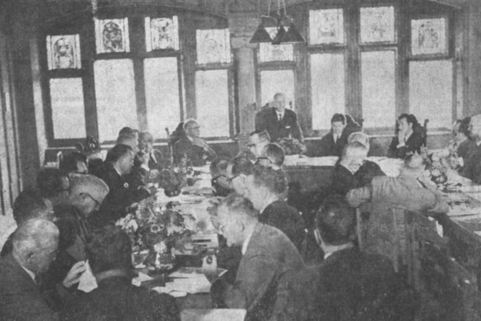 Etwa zwanzig eher ältere Männer sitzen beidseits an Tischen, die in Hufeisenform angeordnet sind. Sie hören einem Redner zu.