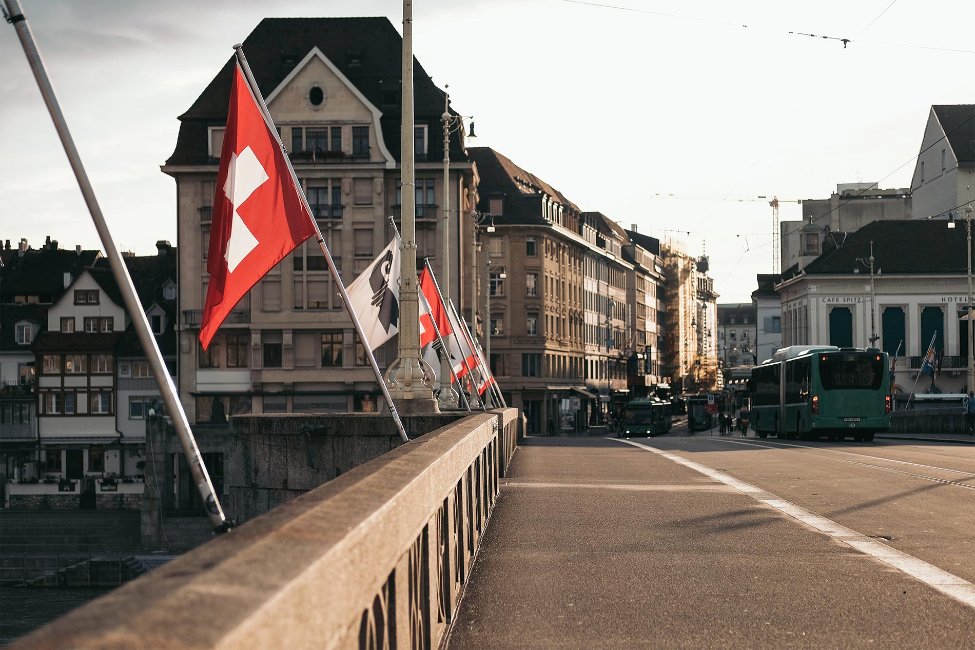 Föderalismus Auf einer Brücke in Basel wehen Fahnen der Schweiz und des Kantons Basel-Stadt im Wind. Im Hintergrund fährt ein Bus in eine Strasse, die von alten Gebäuden gesäumt ist.
